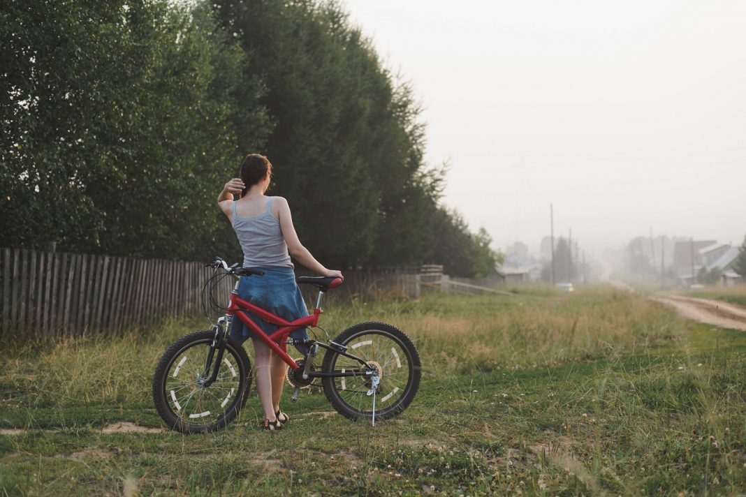 young woman standing with bicycle countryside near garden in village