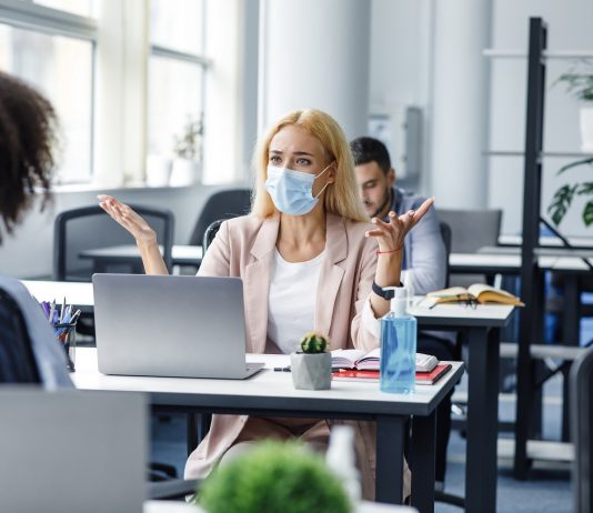 Fogy a türelem, több a vita – mit tehetünk a munkahelyi konfliktusok megelőzéséért? Conflict at work and problems with project. Angry woman in protective mask at workplace with laptop yelling at african american lady and gestures in interior of modern office