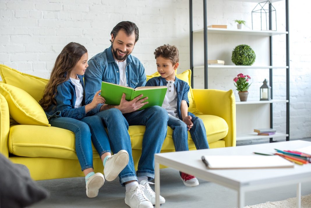 smiling father with children reading book together while sitting on sofa at home