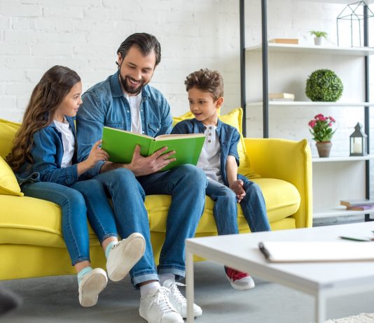 Milyen indokkal és meddig maradhat otthon a szülő a gyermekével a járvány miatt? smiling father with children reading book together while sitting on sofa at home