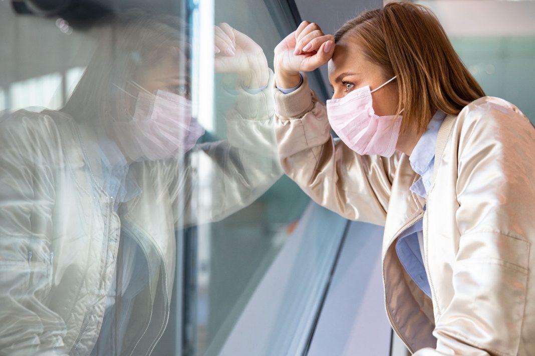 Depressed woman wearing medical face mask, looking out the windo