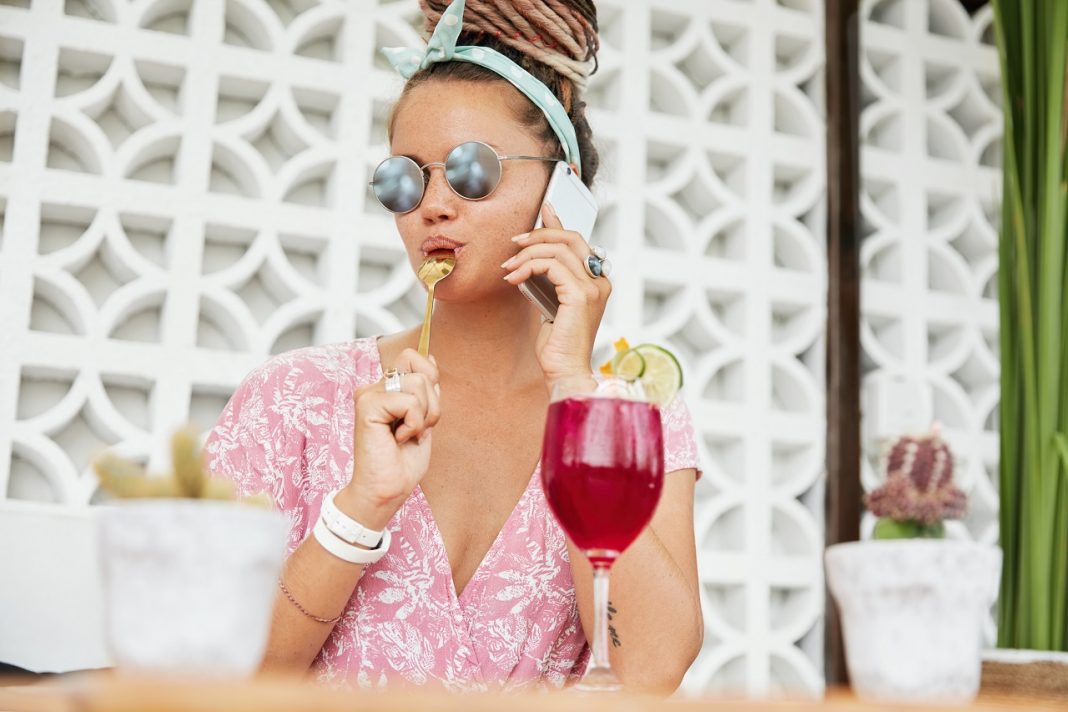 Fashionable young woman in trendy eyewear, licks teaspoon as eats something sweet and drinks cocktail or shake, speaks over modern smart phone, shares her impressions of spent holidays abroad